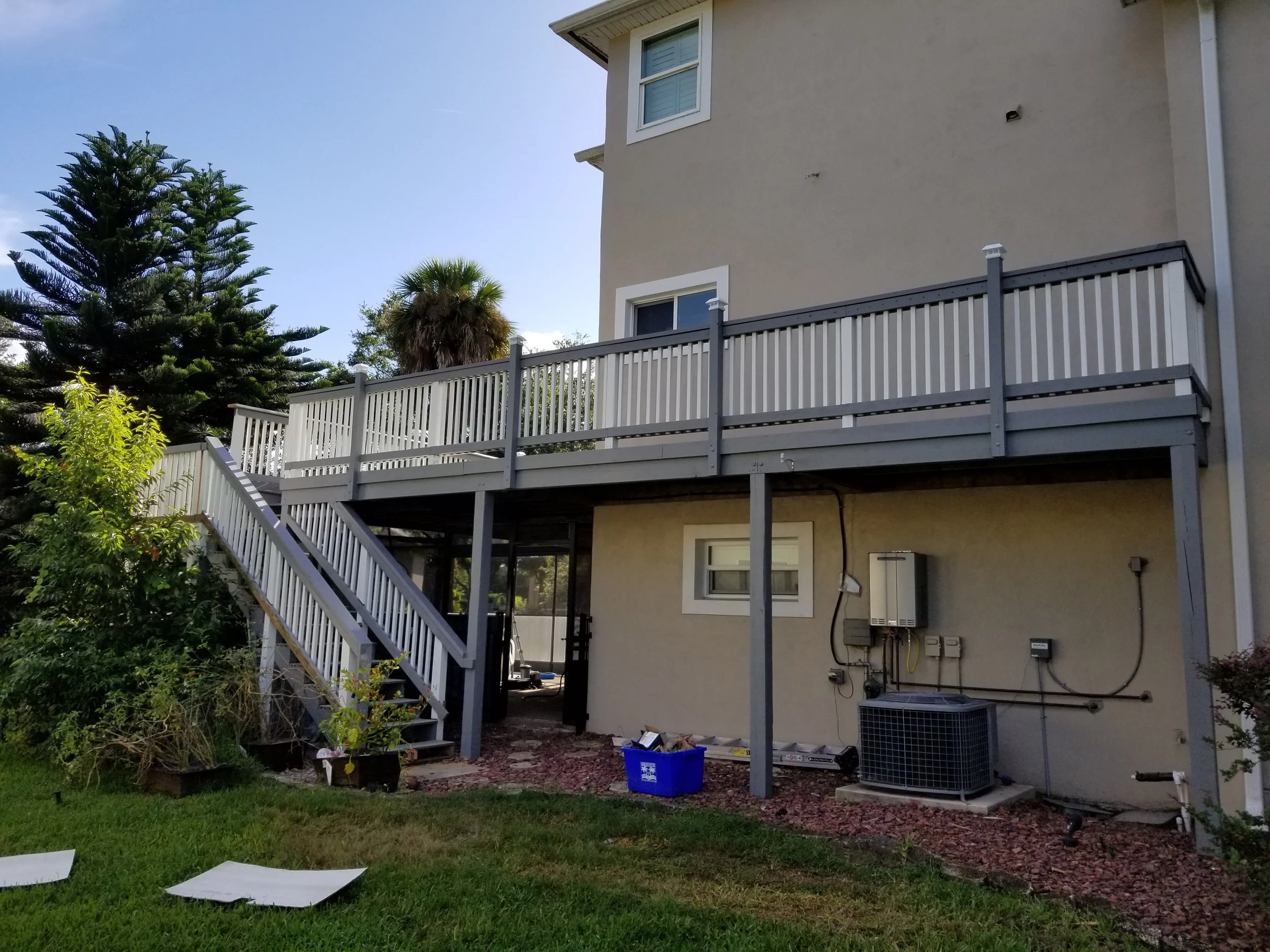 Backyard view of a multi-story house with a white balcony, green trees, and a blue recycling bin. There are some scattered papers or foam pieces on the grass in the foreground.