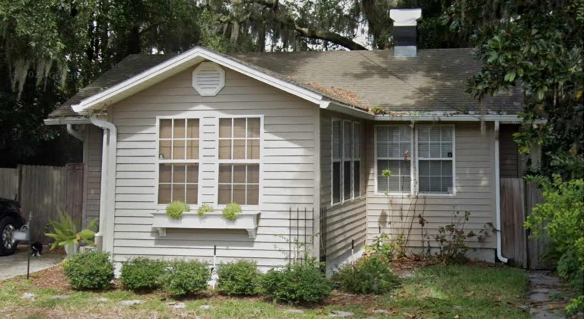 A two-story beige house with multiple windows, a sloped roof with some overgrown leaves, surrounded by a garden with bushes, and trees in the background.