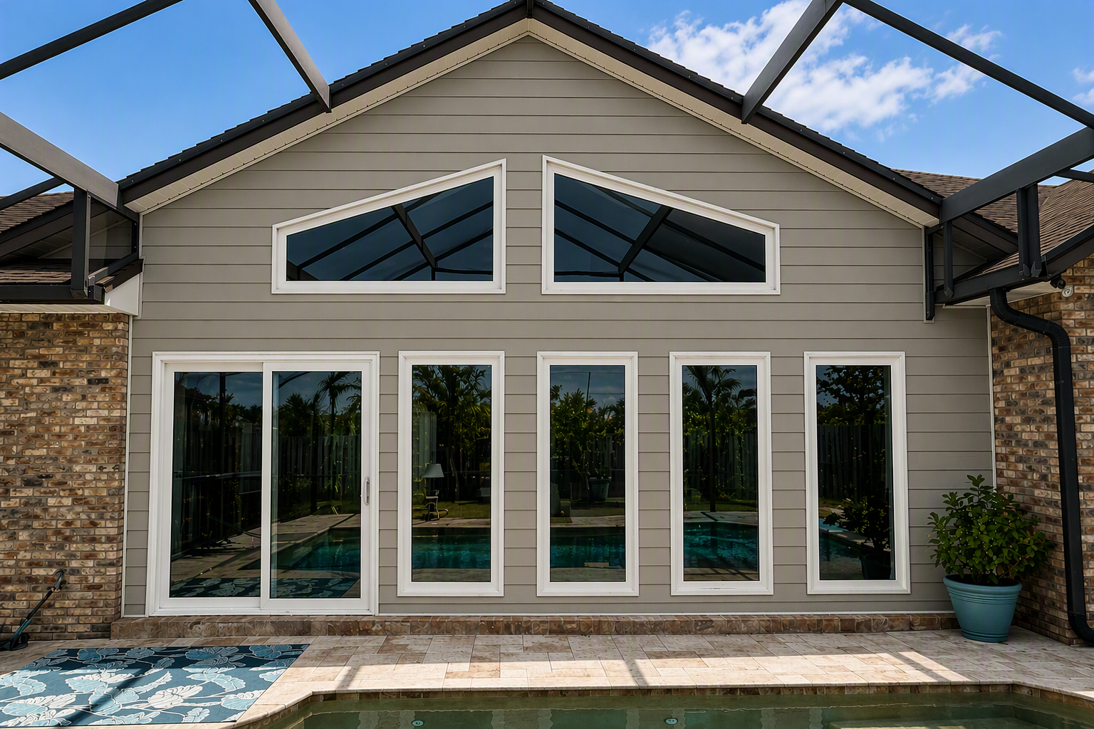 Rear view of a modern house with beige siding, large glass windows, a brick wall on the side, and a backyard with a swimming pool, potted plants, and outdoor furniture. The house has a gabled roof and a screened porch extension.