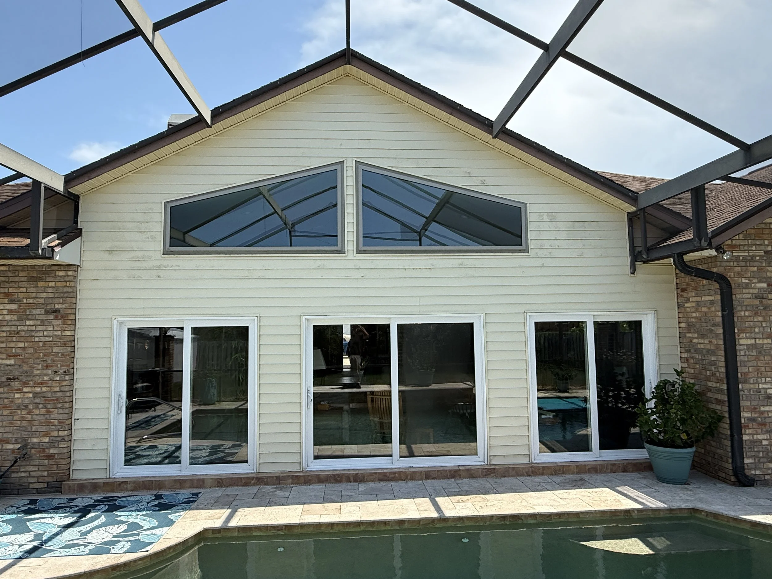 Backyard with a house that has large glass windows and a sliding door, a swimming pool with a tiled deck, and an unfinished metal pergola frame.