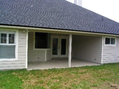 Rear view of a house with a shaded patio, white siding, and small window, next to a grassy yard.