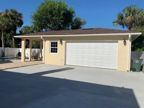 A suburban house with a garage, front porch, and driveway, surrounded by palm trees and greenery on a sunny day.