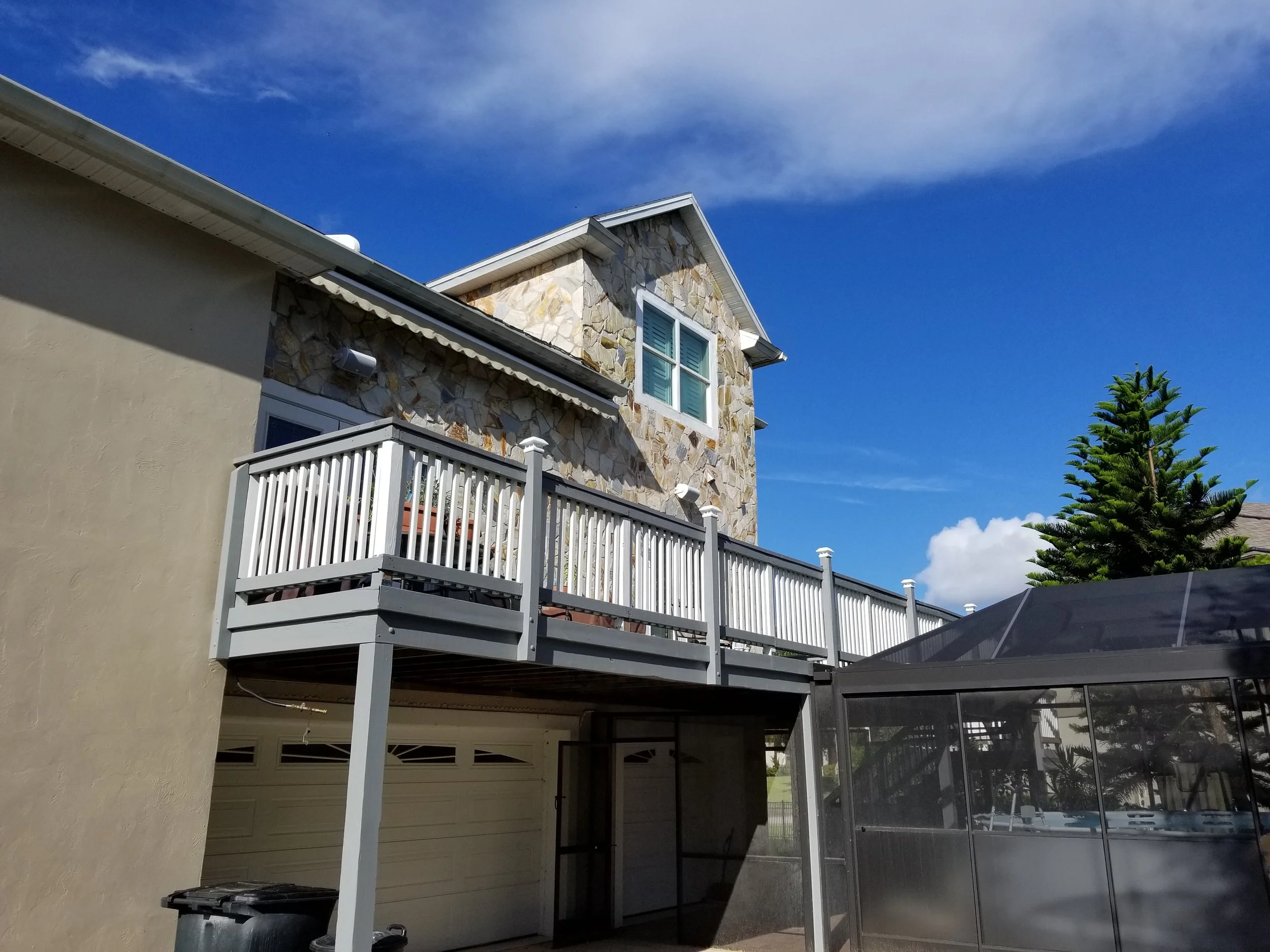 A two-story house with a stone facade and white trim, featuring an upstairs balcony with white railings, a beige stucco wall, a garage door, and a large pine tree in the background under a partly cloudy blue sky.