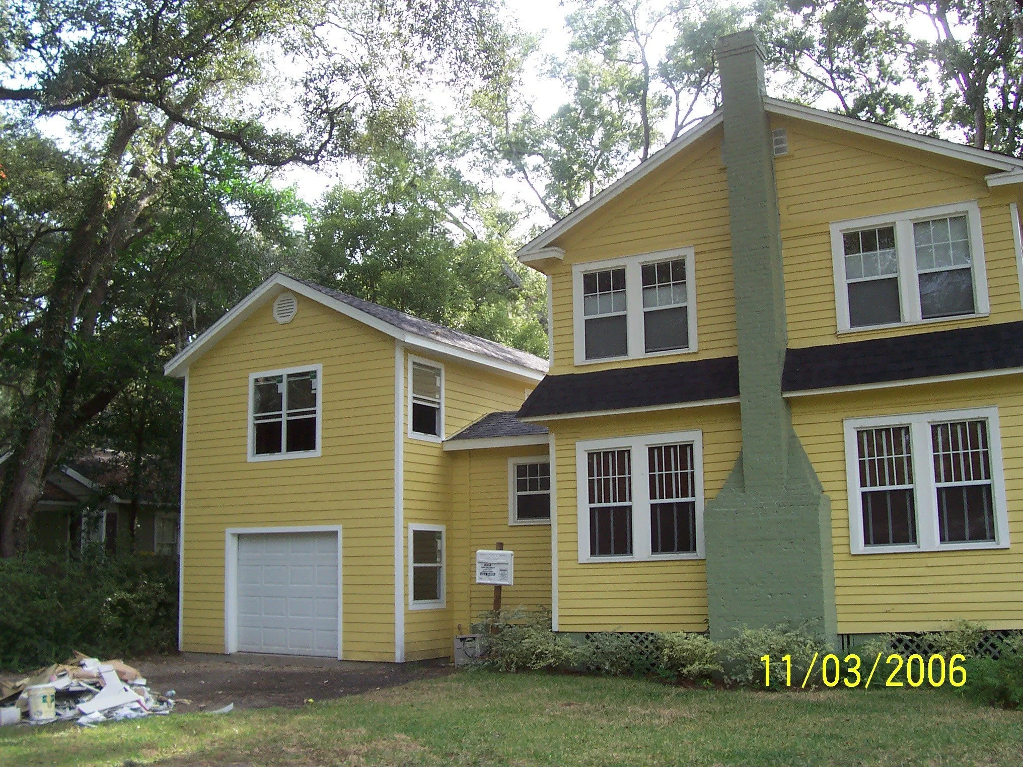 A yellow house with white trim, a chimney, and multiple windows, surrounded by green trees and grass, with a pile of debris in the front yard and a small sign near the entrance, taken on November 3, 2006.