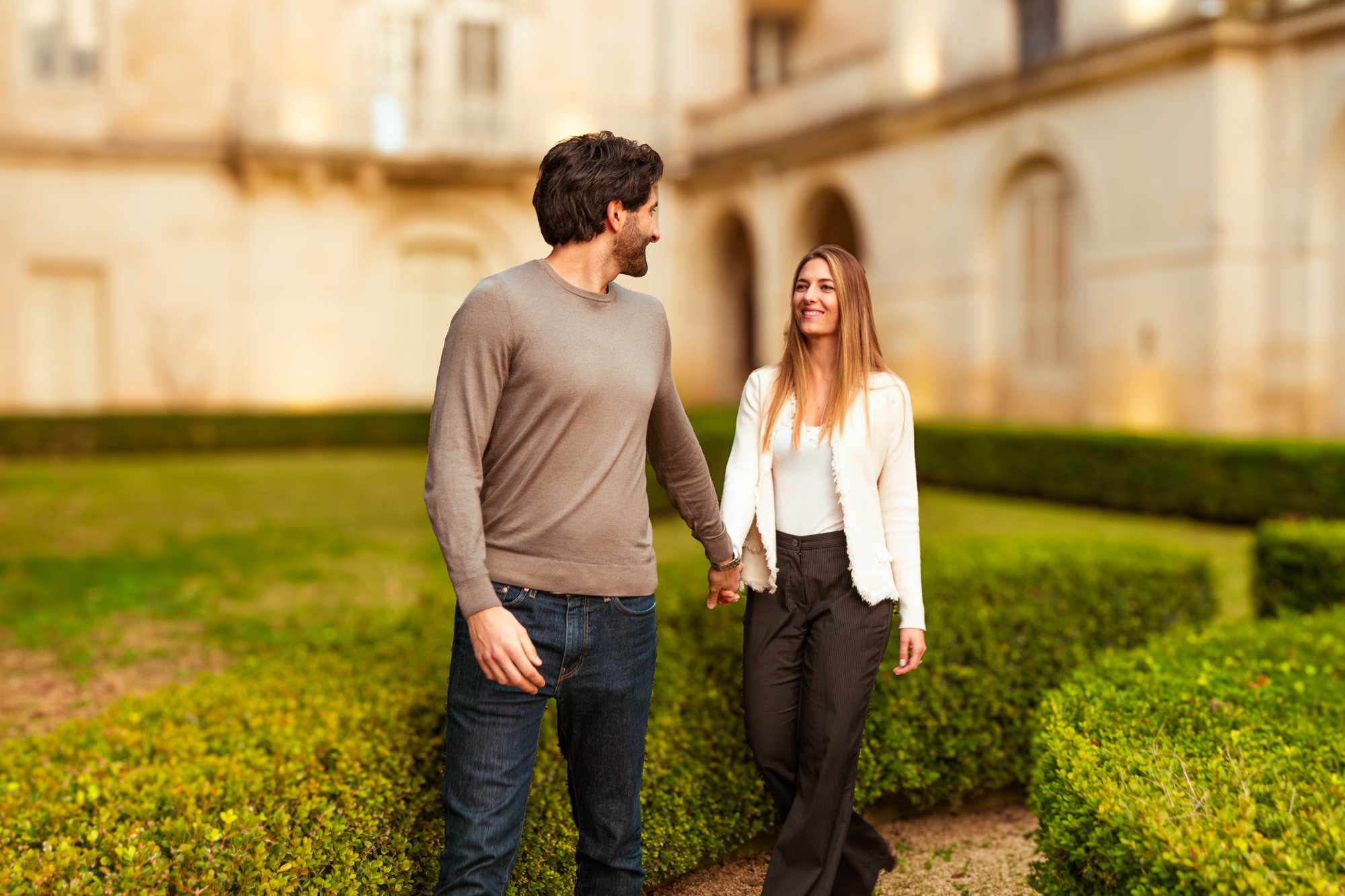 Una coppia si tiene per mano mentre cammina in un giardino con un edificio storico sullo sfondo, sorridendo e guardandosi affettuosamente.