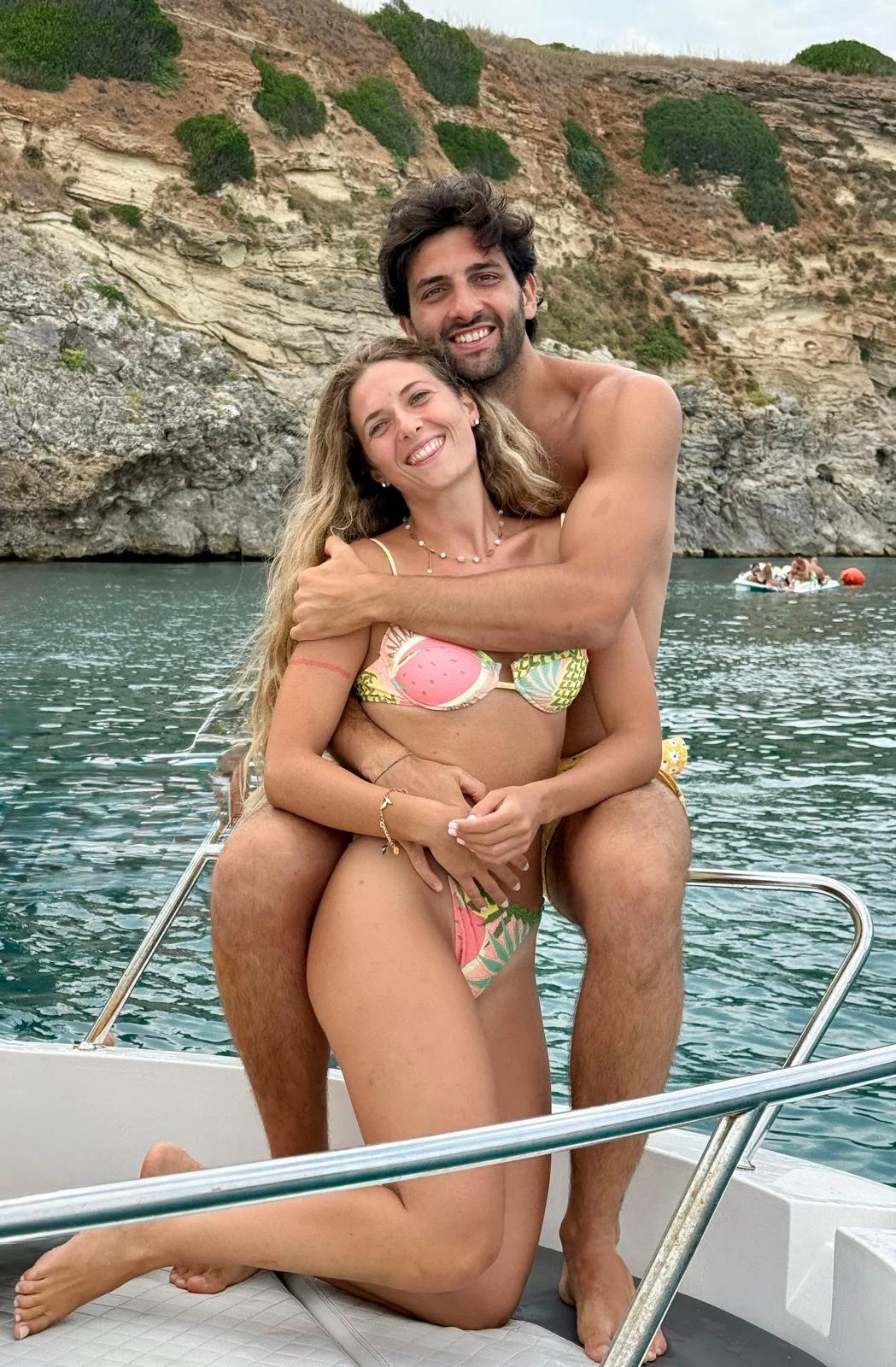 Una coppia di giovani si abbraccia sorridendo su una barca in mezzo al mare, con uno sfondo di scogli e vegetazione, vestiti in costumi da bagno.