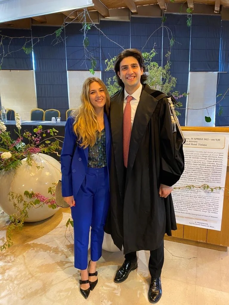 Una giovane donna e un giovane uomo sorridenti, in abiti formali, si abbracciano durante una cerimonia di laurea in un ambiente elegante decorato con piante e fiori.