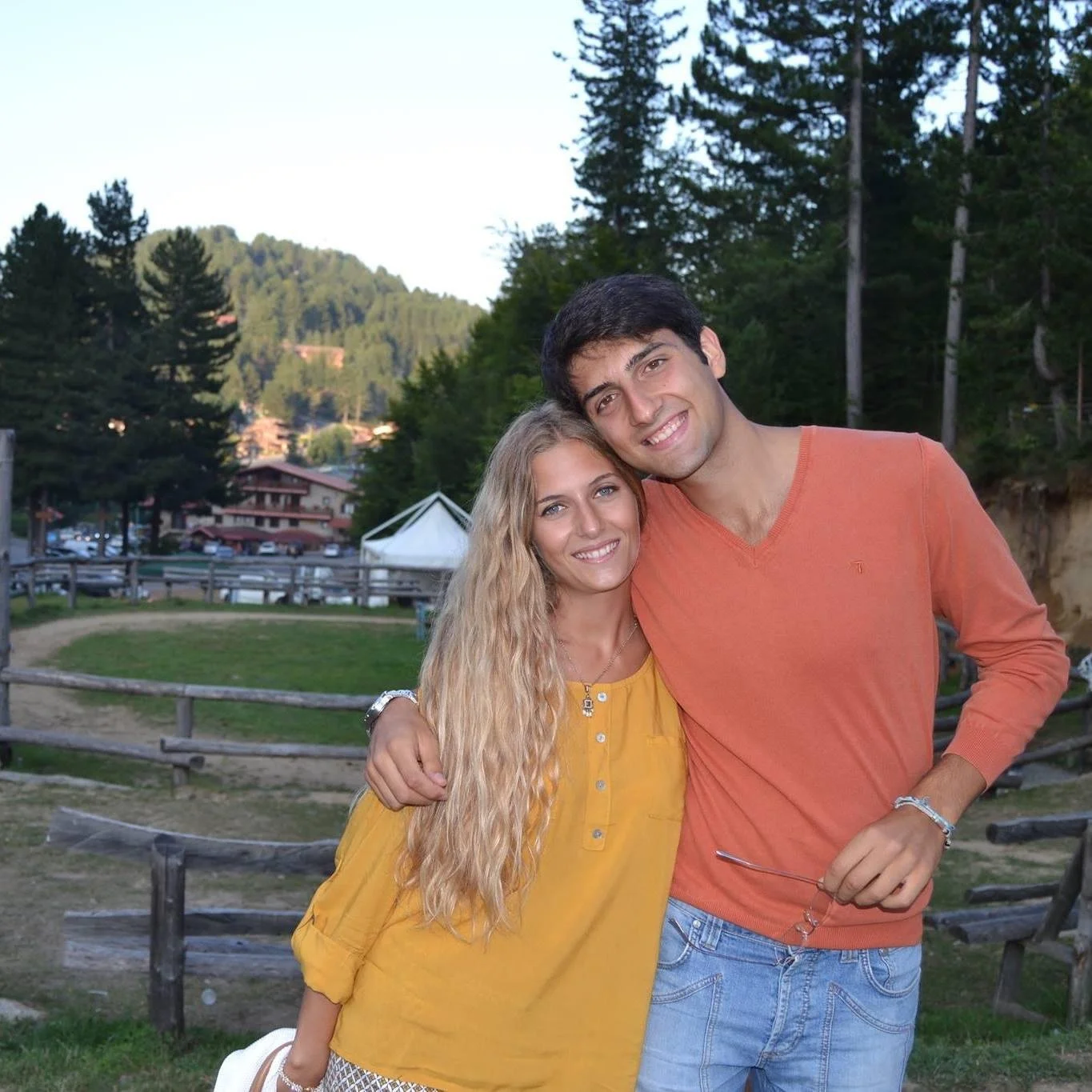 Una coppia sorridente che si abbraccia all'aperto con alberi e edifici sullo sfondo.