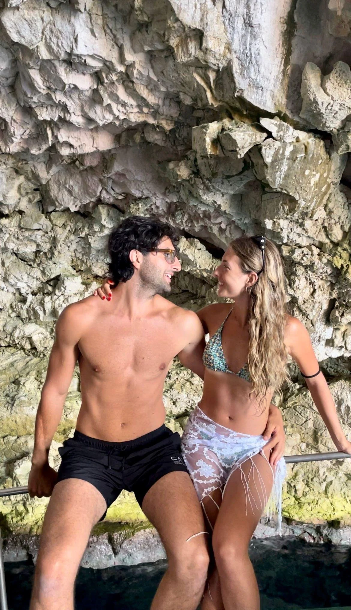 Una coppia di giovani in costume da bagno si siede su una ringhiera vicino a una scogliera, sorridendosi e guardandosi negli occhi.