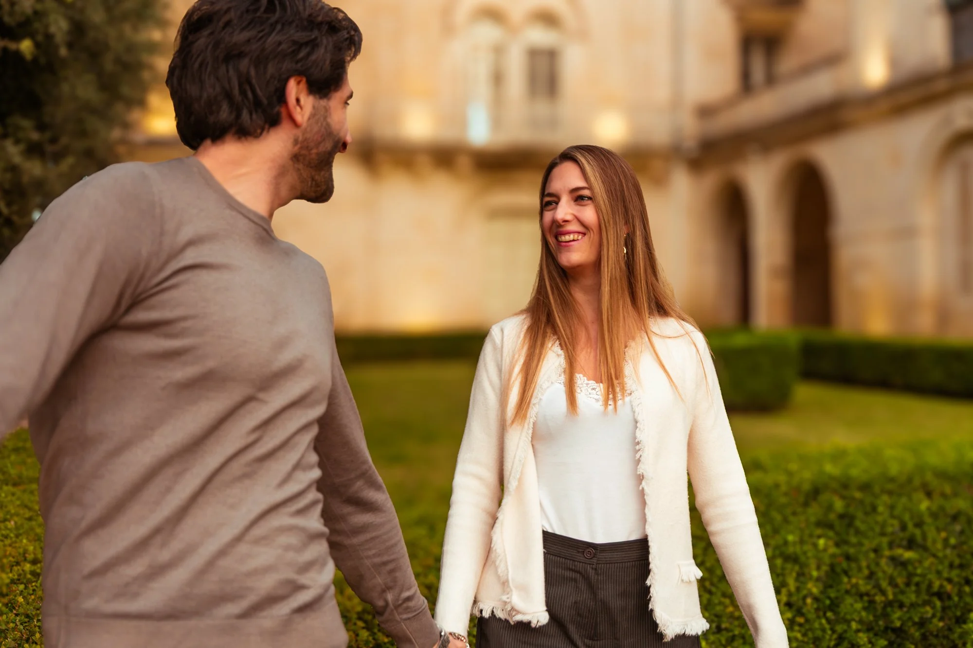 Una coppia sorridente in piedi all'aperto davanti a un edificio storico in pietra durante il tramonto.