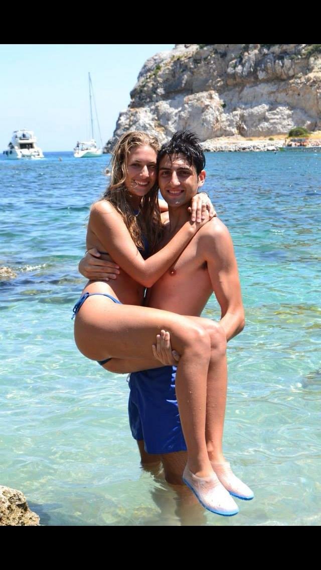 Due persone felici in acqua di mare con una coppia sposata, uomo africano e donna bianca, entrambi abbracciandosi, con barche e scogli sullo sfondo.