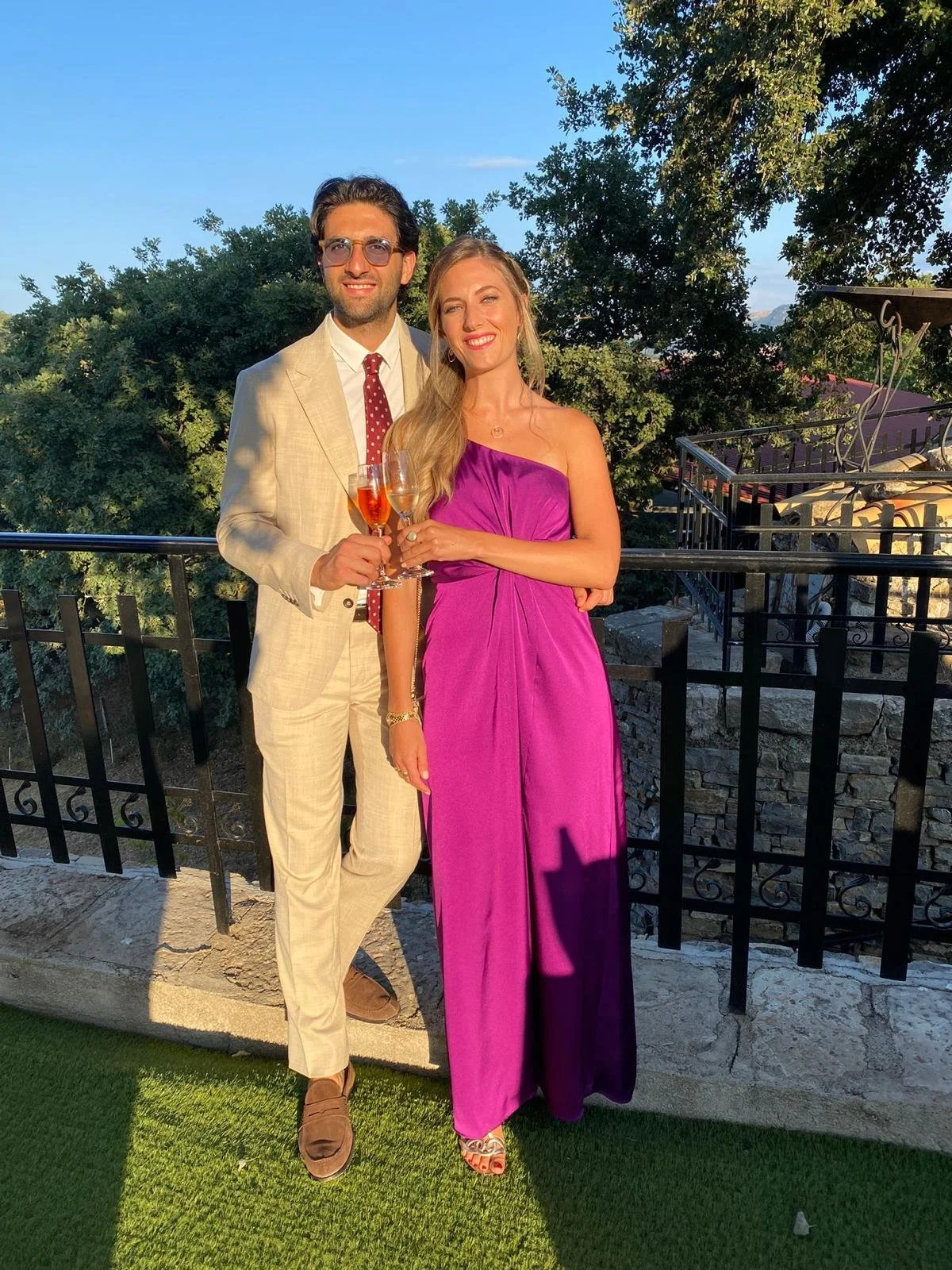 Una coppia elegante che brinda con bicchieri di champagne su un balcone durante il tramonto. La donna indossa un abito lungo rosa e il uomo un completo beige con una camicia bianca e cravatta a pois rossi.