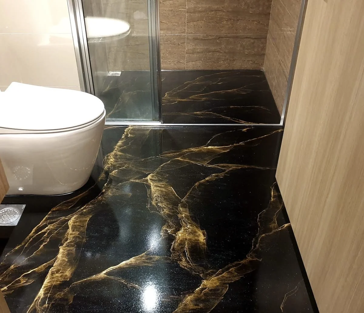Bathroom with black marble floor featuring gold veining, a white toilet, and a glass shower enclosure with brown tiled walls.