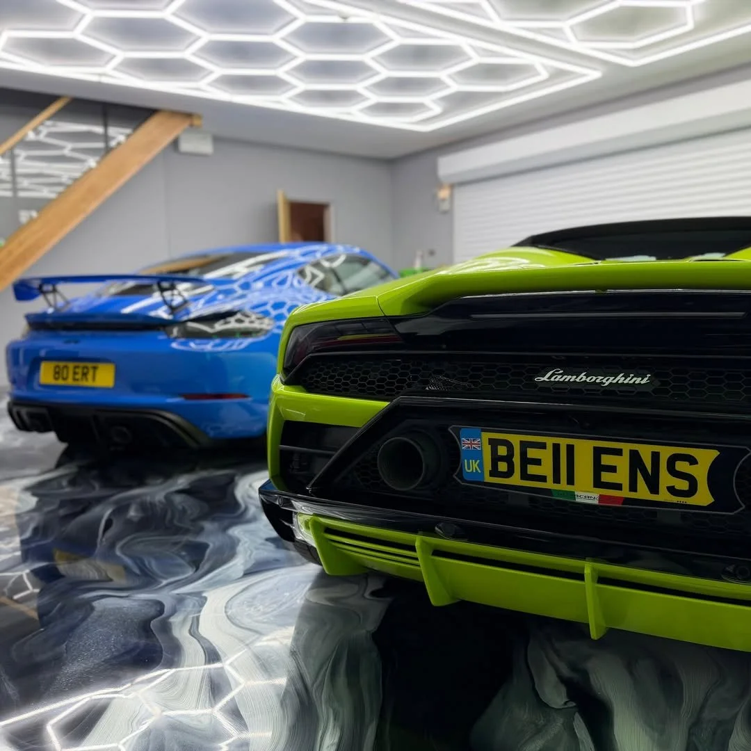 Close-up of a lime green Lamborghini car with a black front grille, in a garage with a blue car in the background, reflective flooring, and modern ceiling lights.