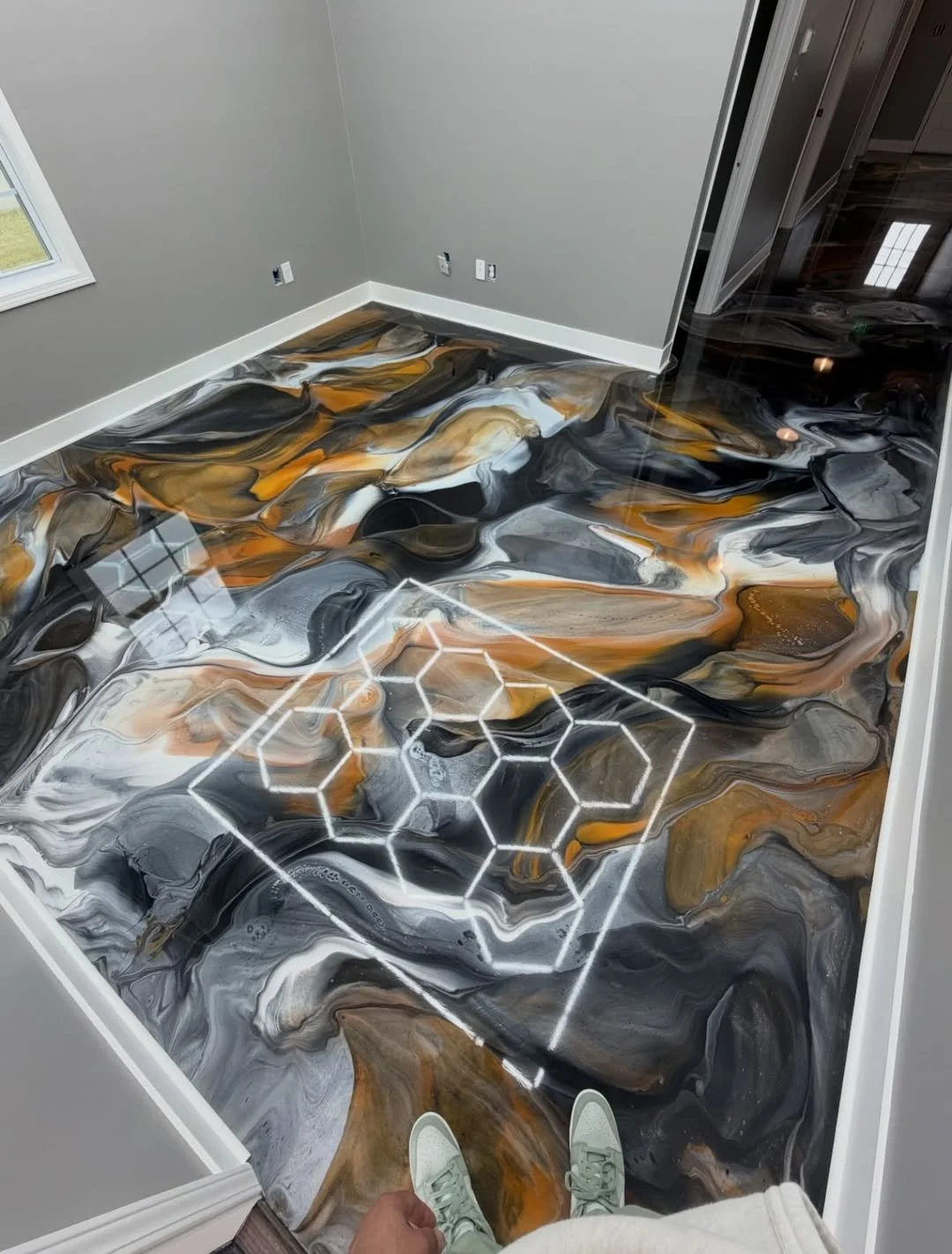 Room with a marble-like black, white, gray, and gold patterned floor and a white geometric design on it, viewed from a person standing near the edge wearing white sneakers.