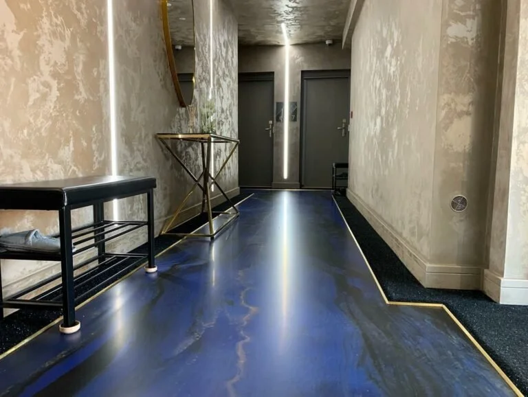 Modern hotel hallway with dark shiny flooring, textured beige walls, a black bench with shoes, a gold and glass table, and two closed elevator doors at the end.