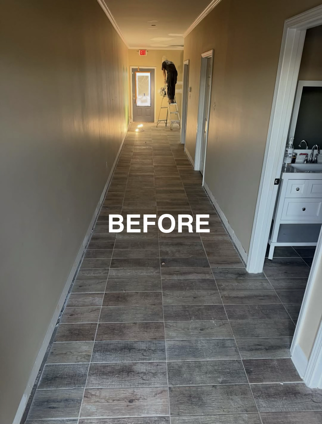 Hallway with beige walls, brown wood-like tiled floor, and a person on a ladder working on the ceiling near the door at the end of the hall.