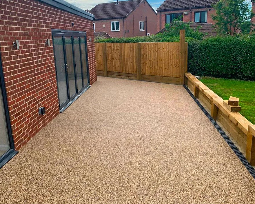 Empty backyard patio with beige textured flooring, a brick house wall with glass sliding doors, a wooden fence at the back, and a small grassy area with hedges and trees.