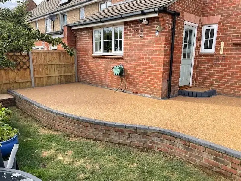 Backyard with newly installed beige textured patio, brick raised border, wooden fence, and brick house with white window and door.