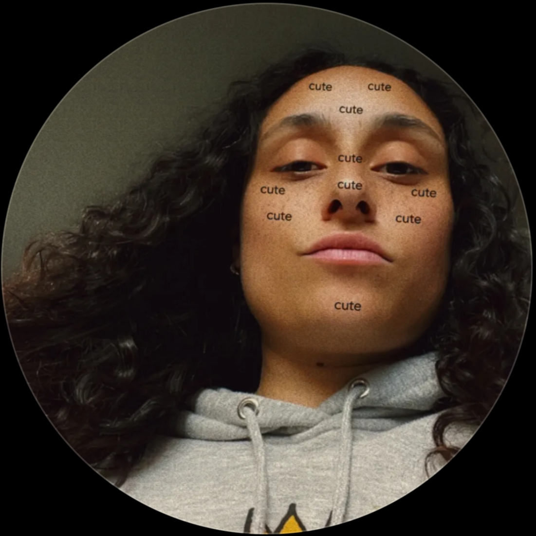 A person with dark curly hair wearing a gray hoodie, with the word 'cute' written repeatedly over their face.