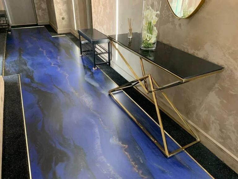 An interior hallway with a glossy blue marble floor, a black console table with a glass vase holding white orchids, and a decorative gold and black console table with angled legs.