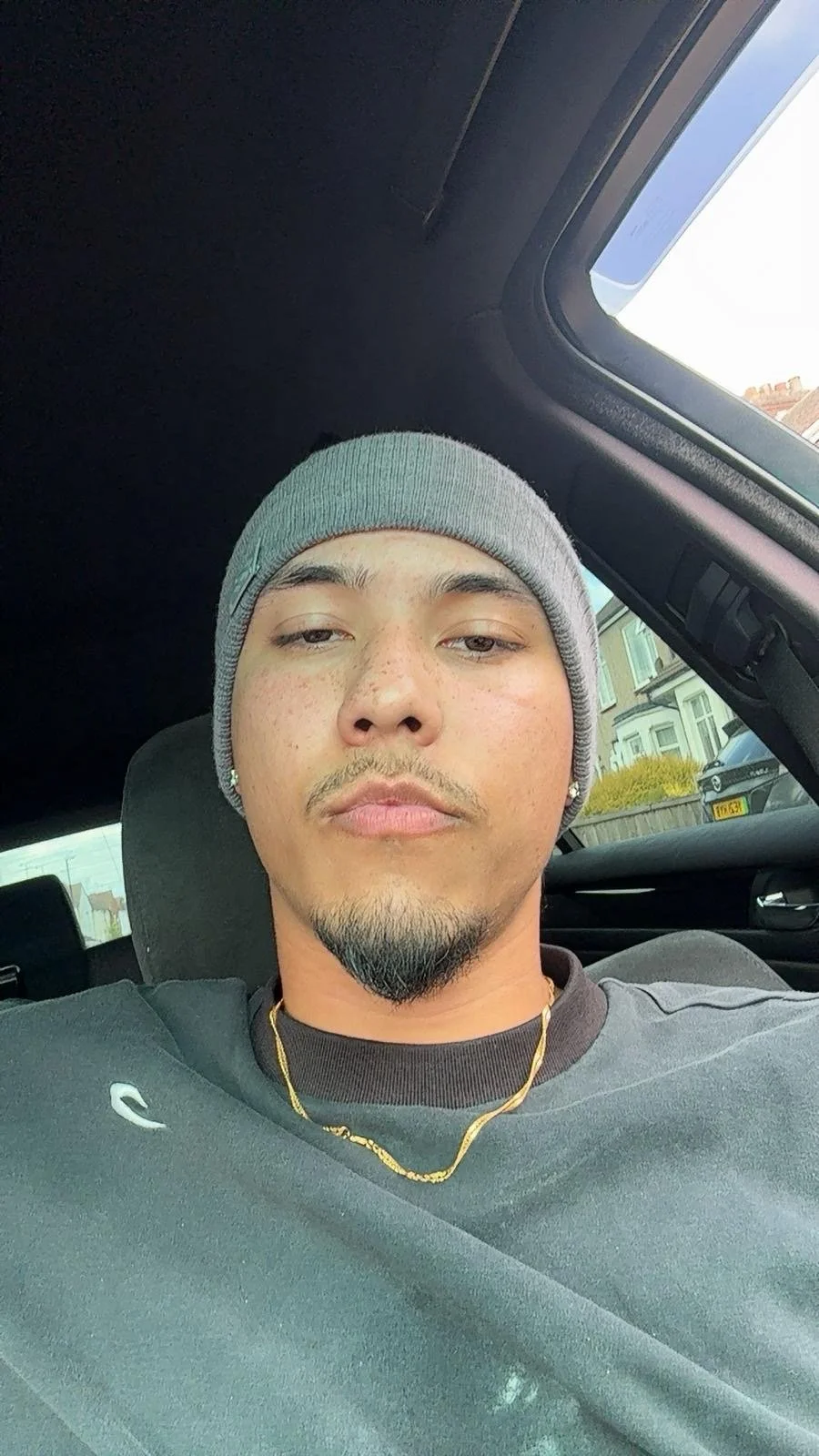 A young man with light brown skin, curly facial hair, wearing a gray beanie, a black shirt, gold chains, and earrings, sitting in a car, taking a selfie.