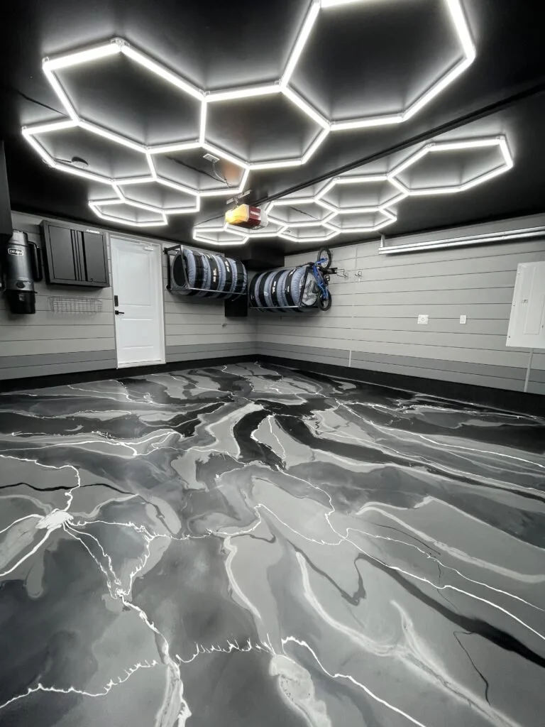 Modern garage with hexagonal LED light ceiling design, marble-patterned epoxy floor, wall-mounted storage racks with tires and a bicycle, gray shiplap walls, and a white door.