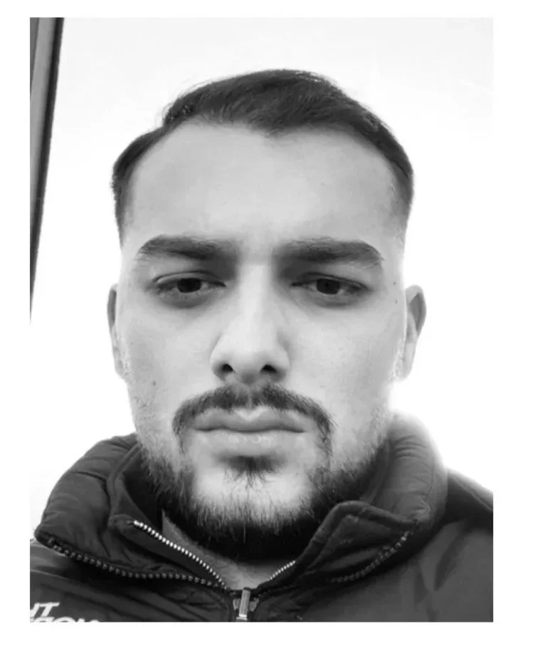 Black and white close-up portrait of a young man with short hair, beard, and mustache, wearing a zip-up jacket with the collar up.