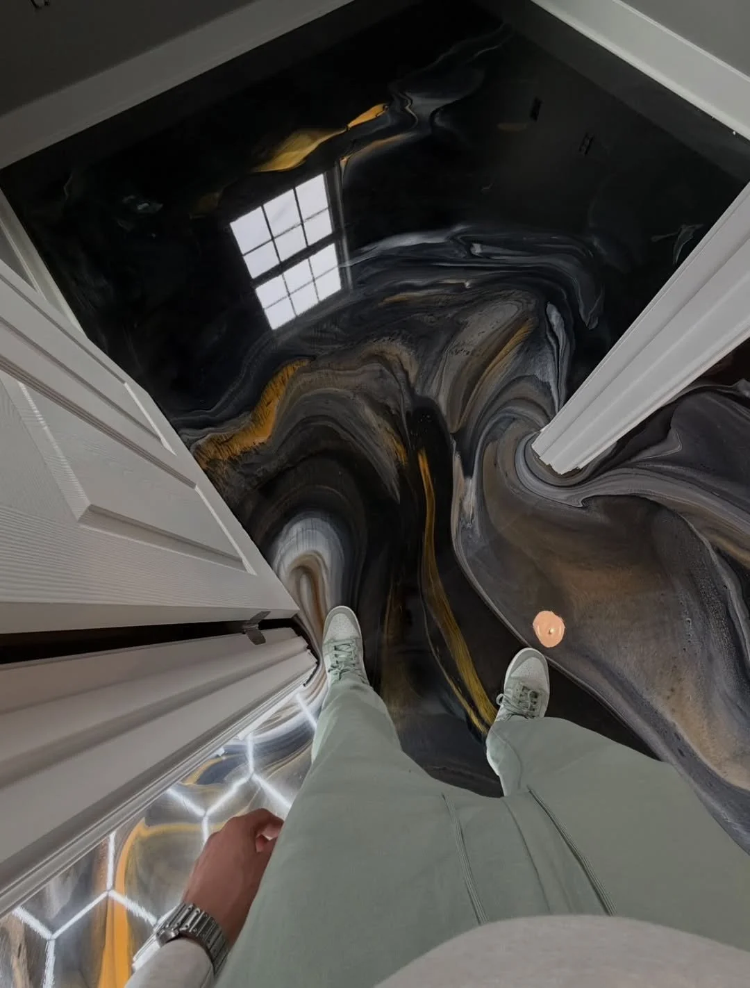Photo looking downward at a person's legs and feet standing on a swirling black and gold marble floor with a window reflection in the background.