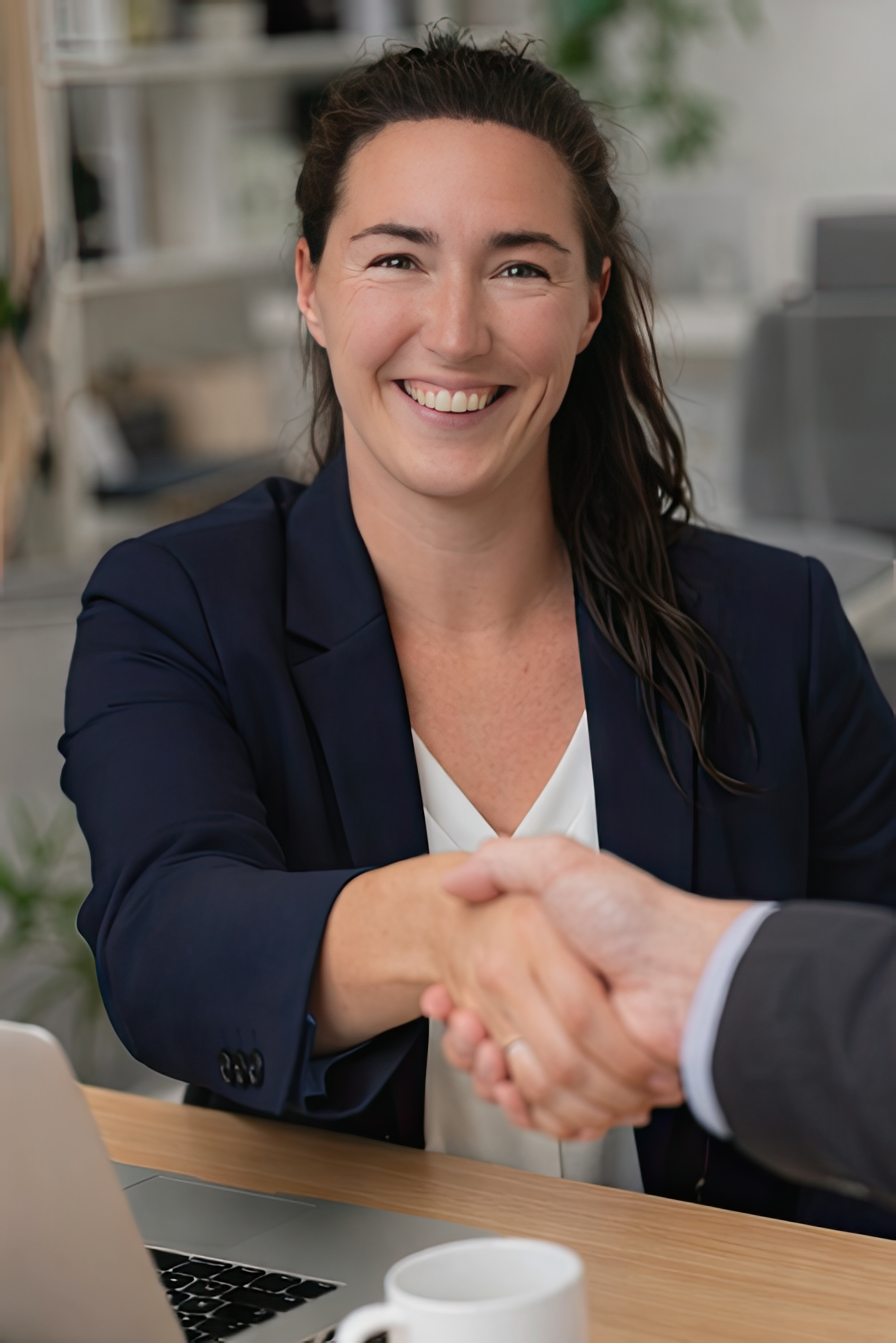 A woman in a navy blazer smiling while shaking hands in an office setting.