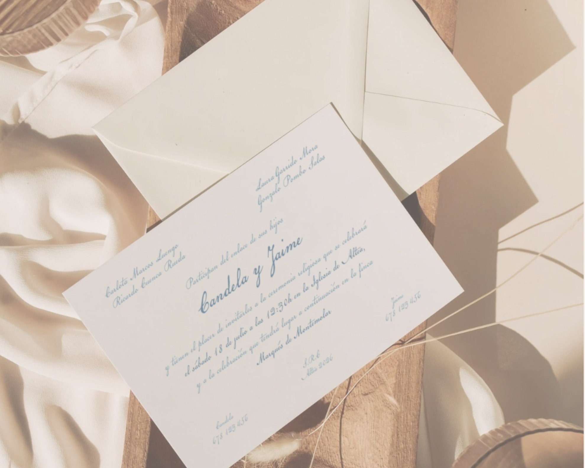 Invitación de boda en color azul, con sobre blanco, rodeada de telas blancas y elementos decorativos de madera.