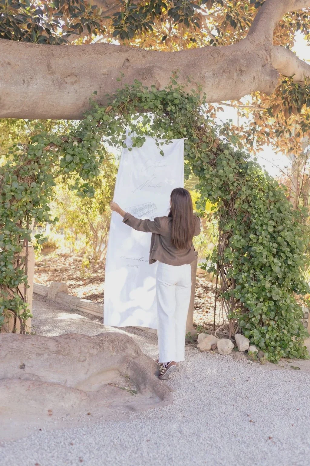 Una mujer de espaldas, de cabello largo, con chaqueta marrón y pantalones blancos, levanta una tela grande en forma de pantalla en un arco cubierto de vegetación, en un entorno al aire libre con árboles y suelo de grava.