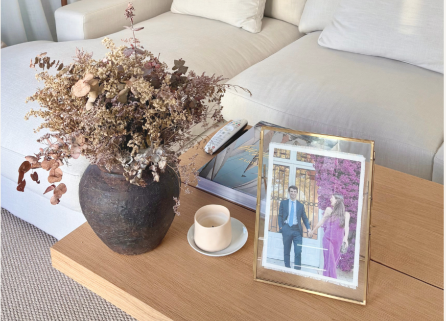 Vaso con flores secas, una fotografía enmarcada de una pareja, un libro, un control remoto y una vela en una mesa de madera en una sala de estar con sofá blanco.