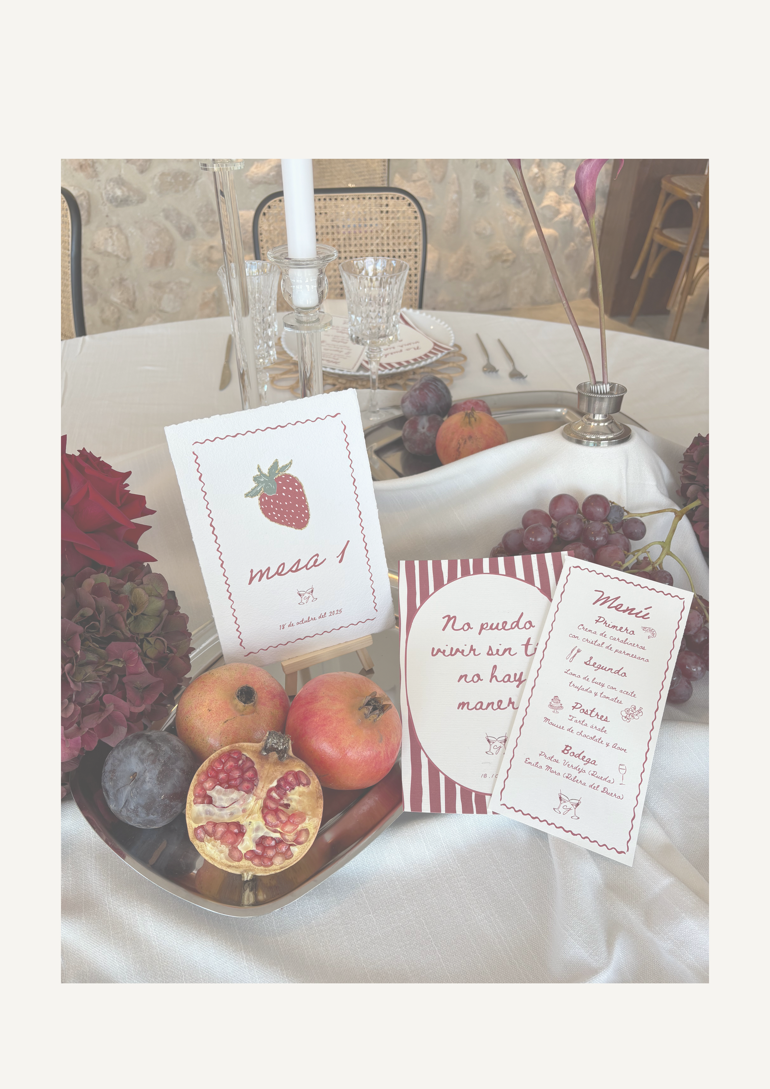 Mesero con un plato de frutas y cartas en una mesa decorada con flores rojas y uvas, en un entorno elegante con velas y cristalería.