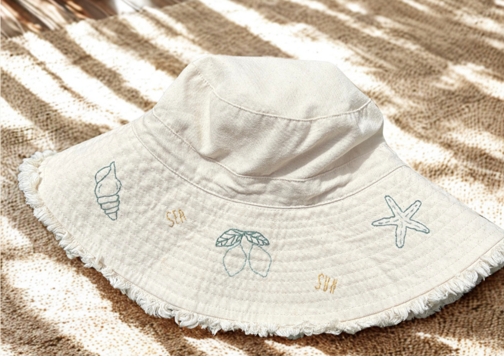 Sombrero de playa blanco con bordados de concha, estrella de mar, hoja y la palabra 'SUN' en colores claros, colocado sobre una toalla de rayas marrón y beige.