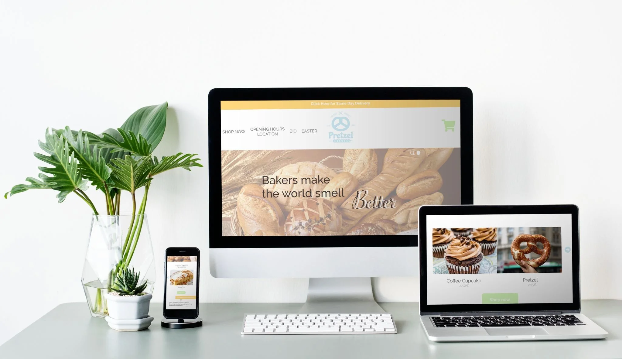 Arbeitsplatz mit einem iMac, einem MacBook, einem iPhone, einem Holz Tisch, einem grünen Pflanzen Topf, und einer Pflanzen. Der Bildschirm des iMac zeigt eine Webseite über Bäckereiwaren, mit dem Text "Bakers make the world smell better".