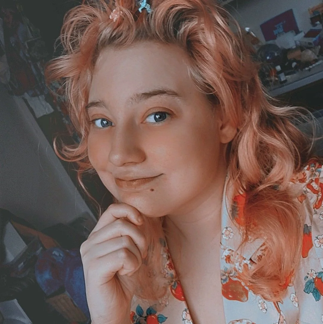 Junge Frau mit hellrotem, lockigem Haar und blauen Augen, die die Hand unter dem Kinn hält, vor unscharbem Hintergrund mit Möbeln und Gegenständen.