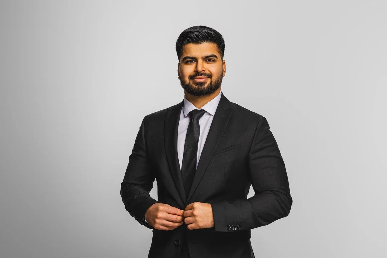 Ein mann in einem schwarzen Anzug, weißes Hemd und schwarzer Krawatte steht vor einem grauen Hintergrund.