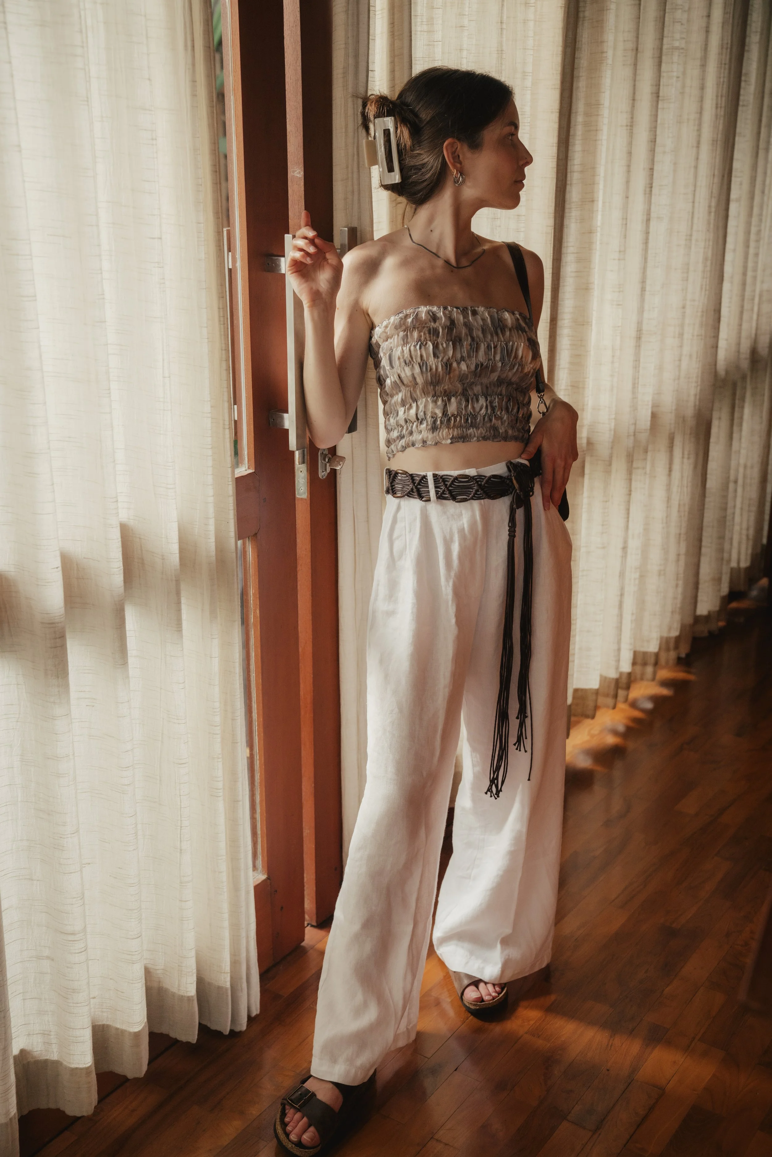 A woman standing indoors near a window with closed curtains, wearing a strapless ruched top and wide-leg beige pants with a black woven belt, showcasing a casual yet stylish outfit.