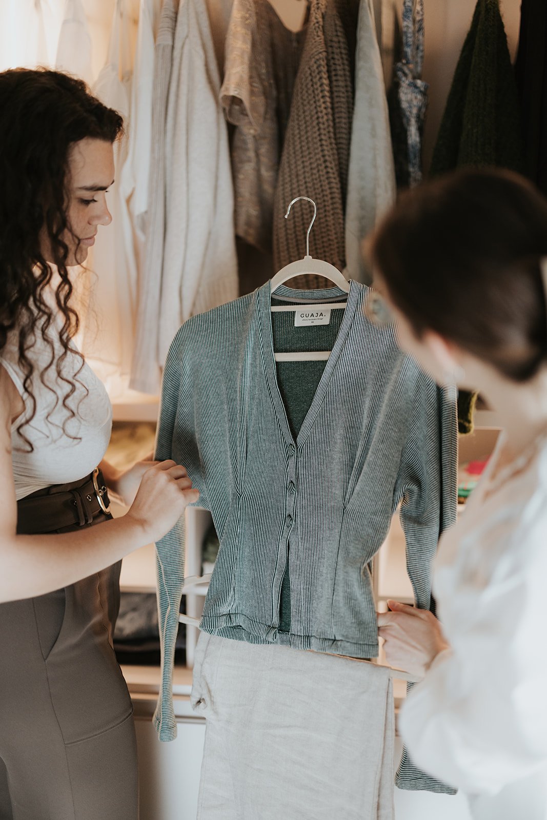 Two women are examining a striped gray cardigan hanging on a white hanger in a closet, with various other clothes hanging behind them.