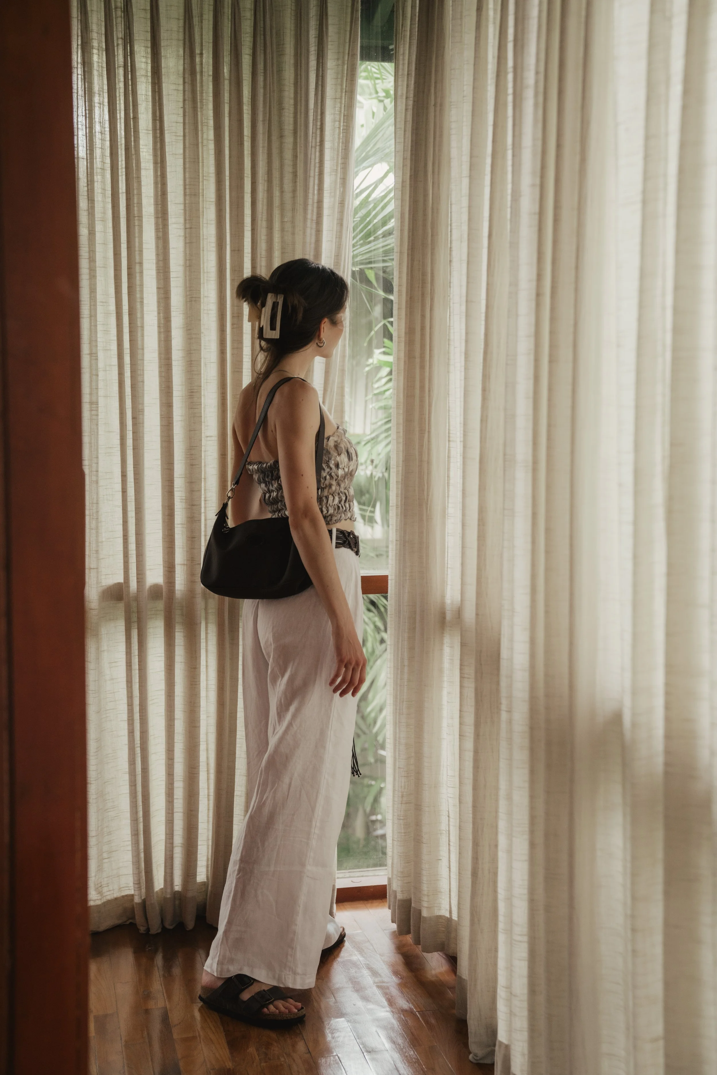 A woman stands in front of a window with beige curtains, looking outside. She wears a sleeveless top, white wide-leg pants, black sandals, and has a black shoulder bag. Her hair is styled with a large clip.