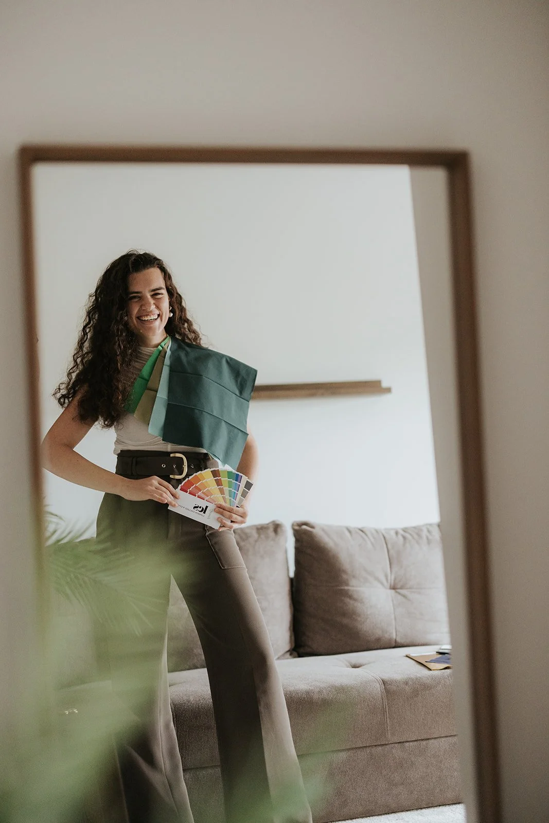 Woman smiling and holding a color swatch fan, standing in front of a mirror, with fabric samples draped over her shoulder in a living room with a beige couch.