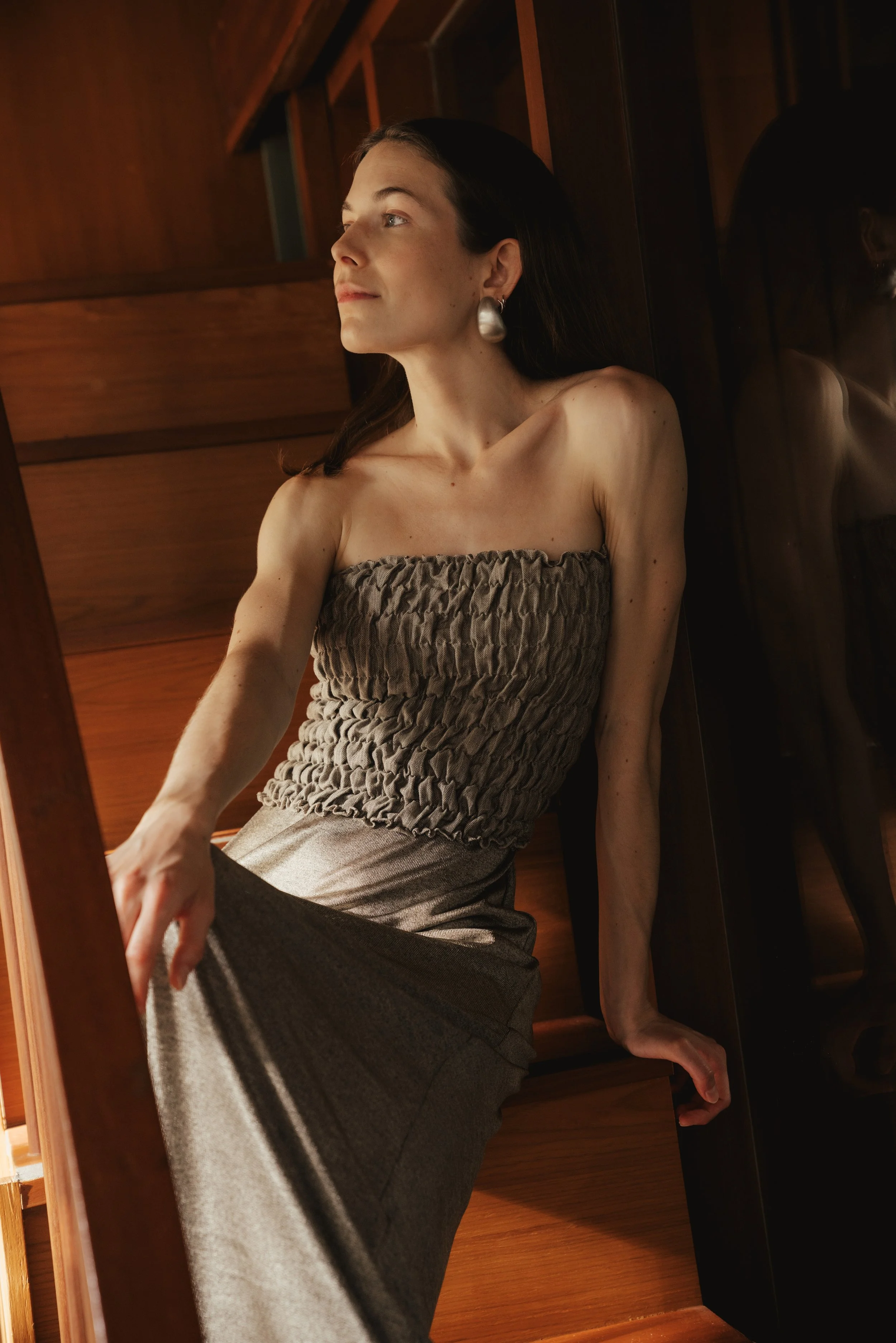 A woman with dark hair wearing a strapless smocked top and a long skirt, sitting against a wooden wall and looking to her left.