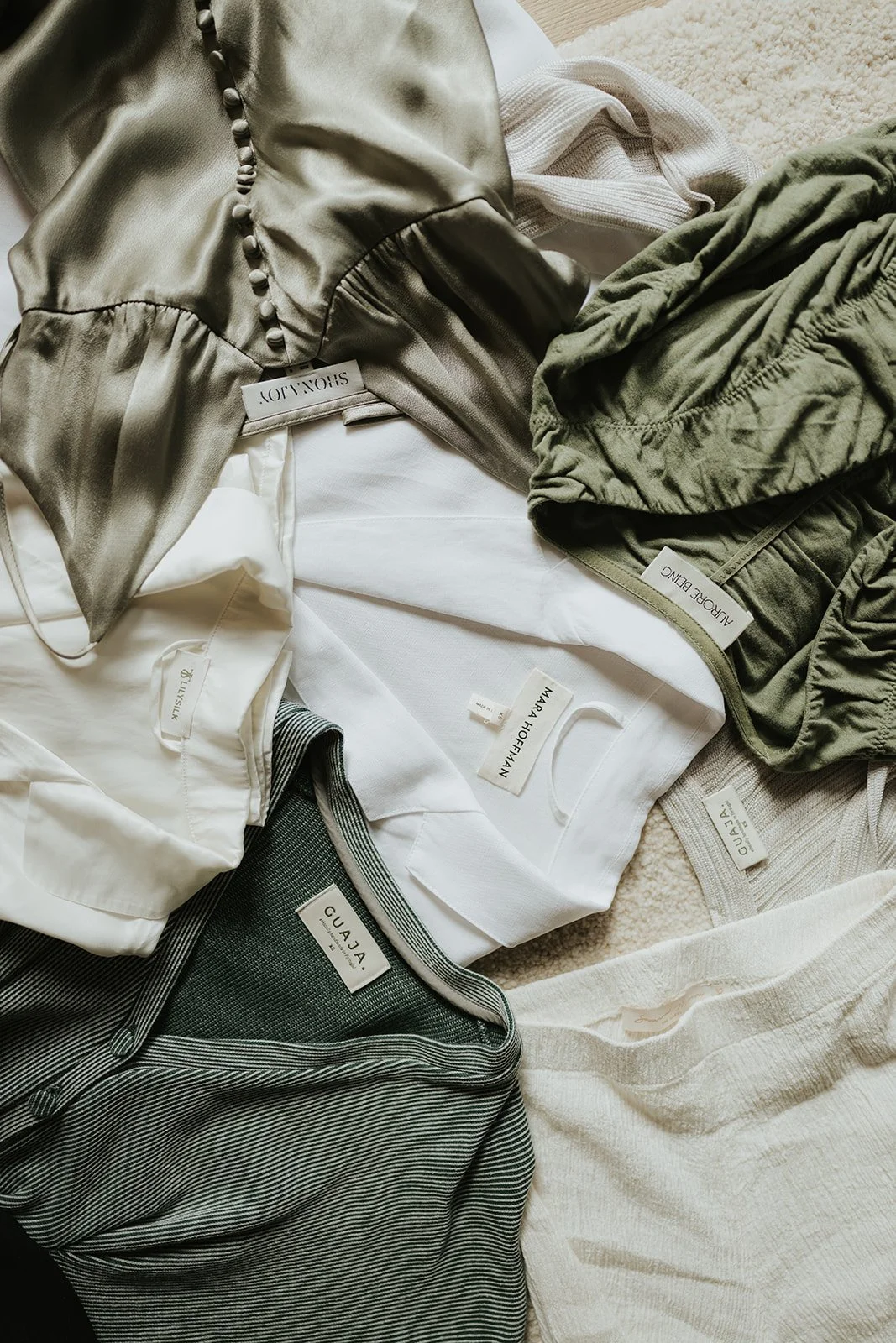 A flat lay of various clothing items including a satin top, a white shirt, green shorts, gray striped pants, and other garments with visible brand tags.