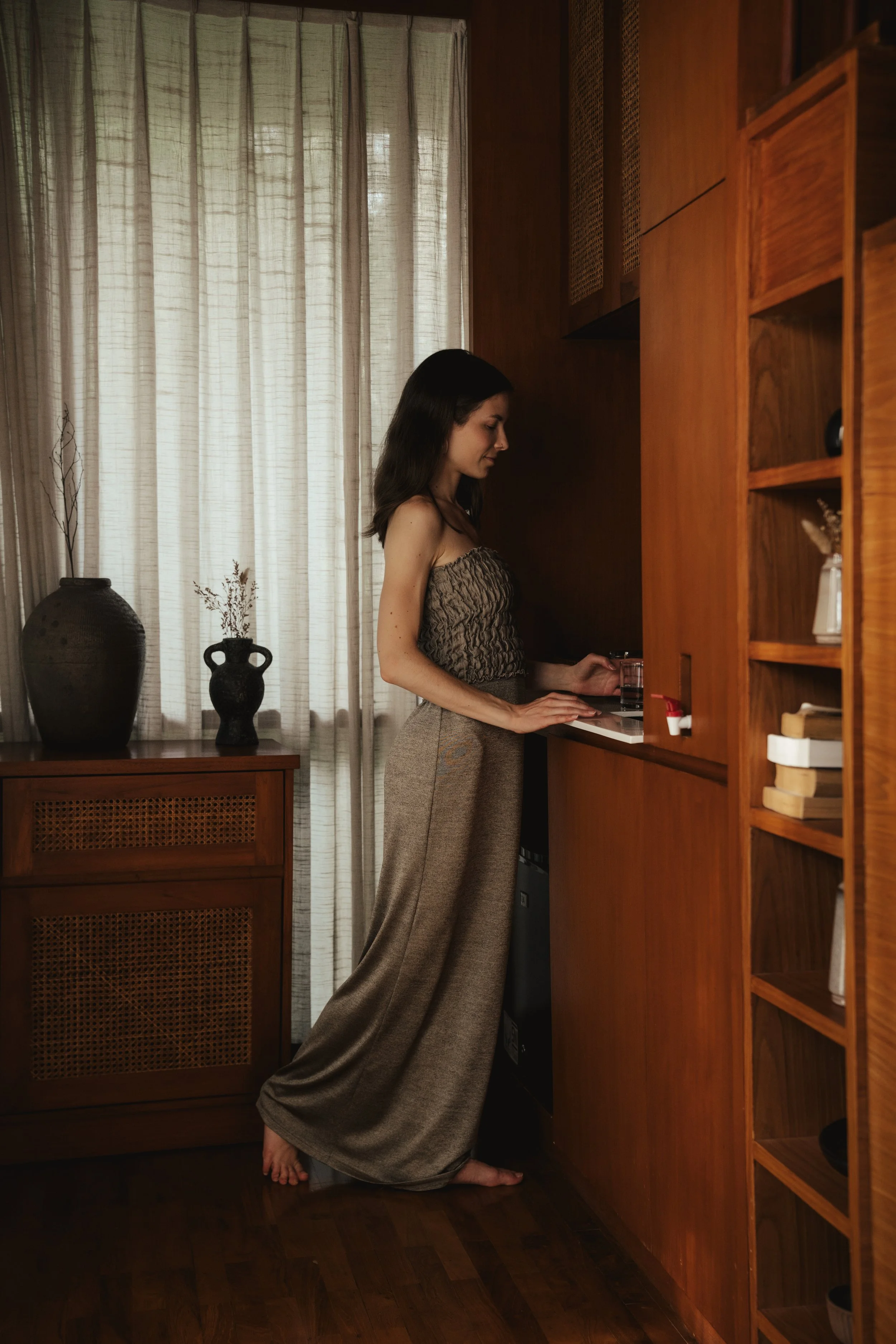 A woman with dark hair, wearing a strapless patterned top and loose beige pants, stands barefoot in a cozy room near a wooden counter, looking down at a small glass of water.