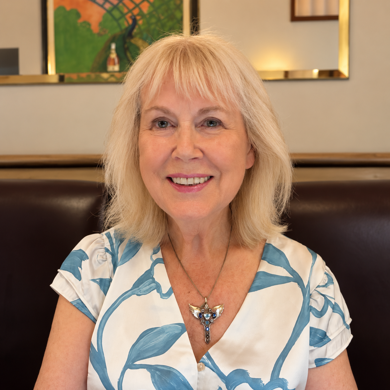 A smiling middle-aged woman with blonde hair, wearing a white blouse with blue leaf patterns and a necklace with a cross-shaped pendant, sitting in a restaurant with framed artwork on the wall behind her.