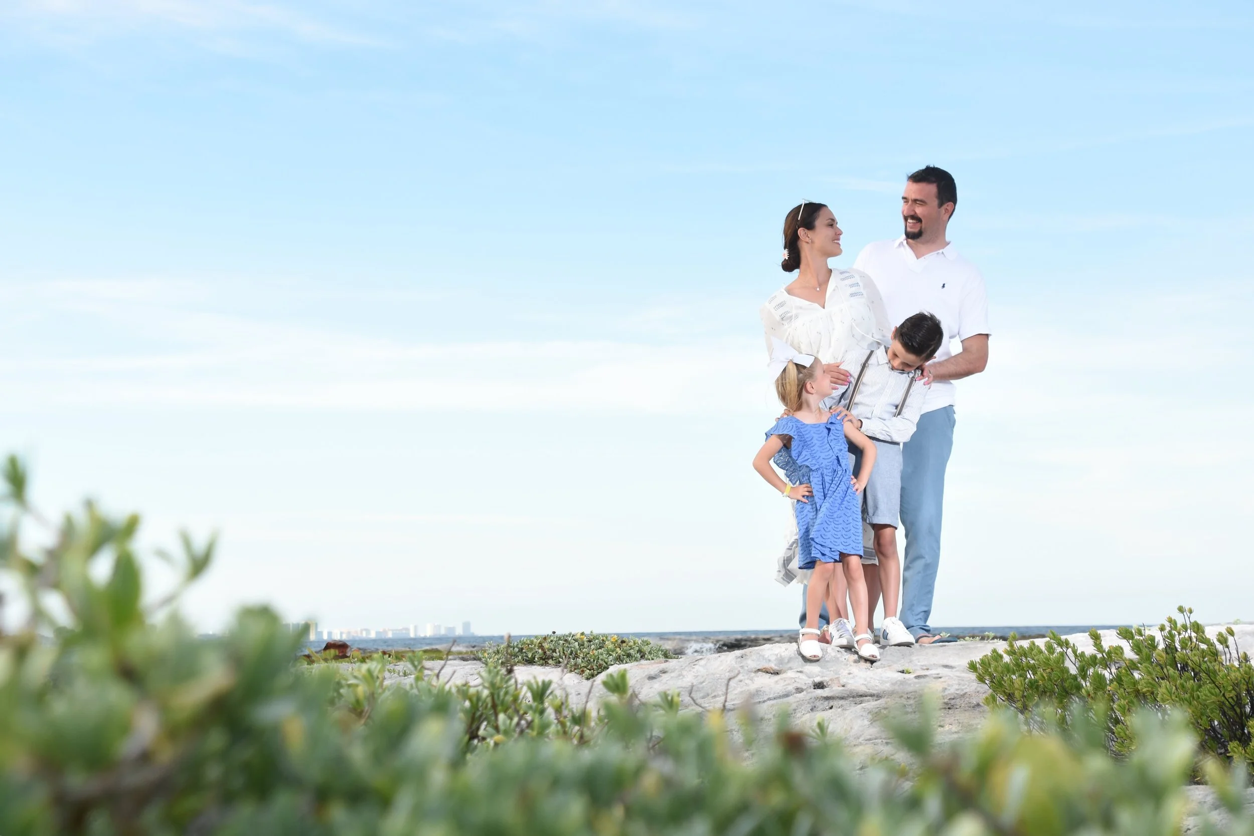 Family of four standing on a rocky beach, smiling, with the ocean and city skyline in the background, under a blue sky.