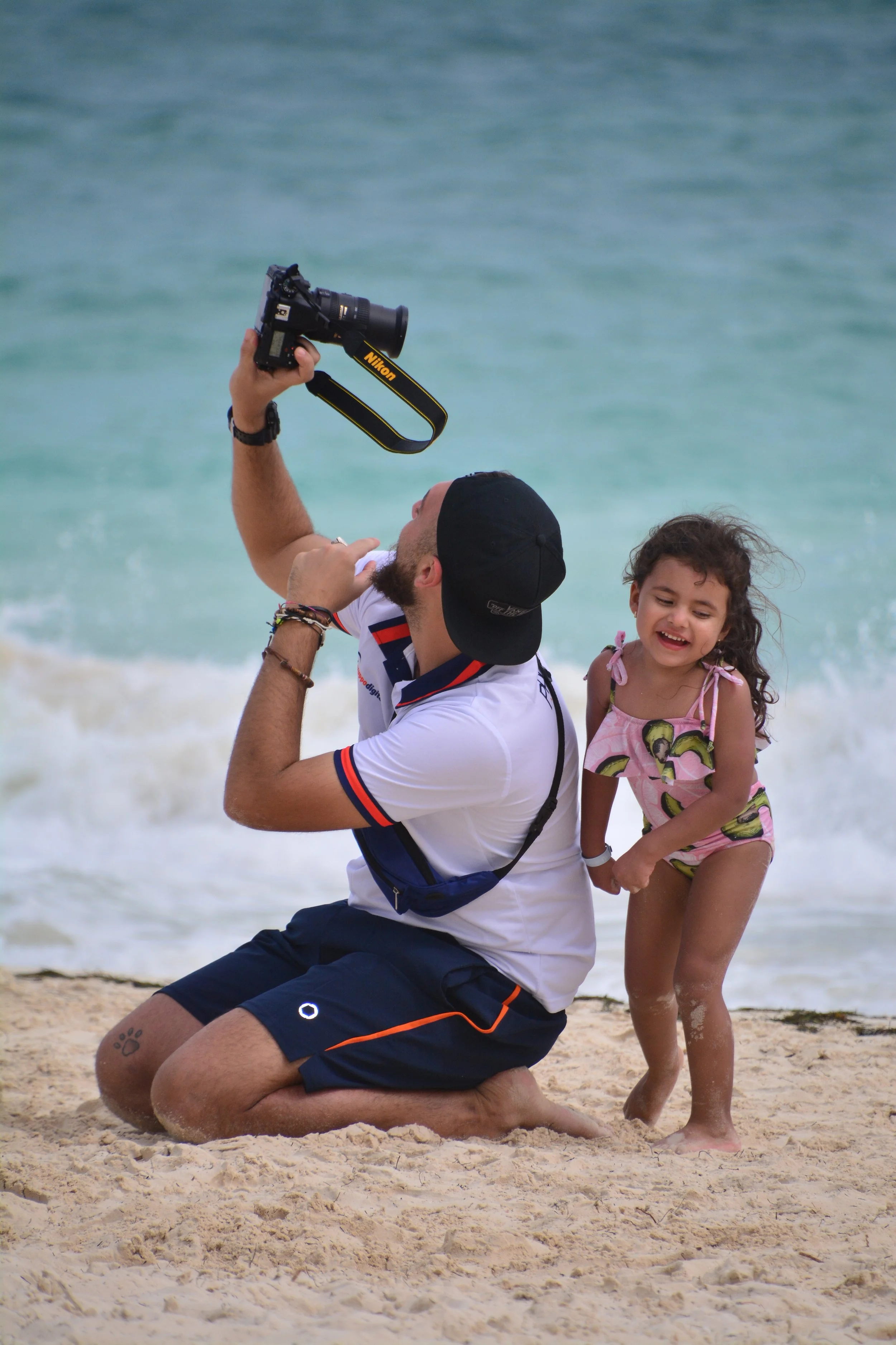 A man kneels on the beach taking a photo with a Nikon camera, while a young girl in a pink and green swimsuit laughs beside him.