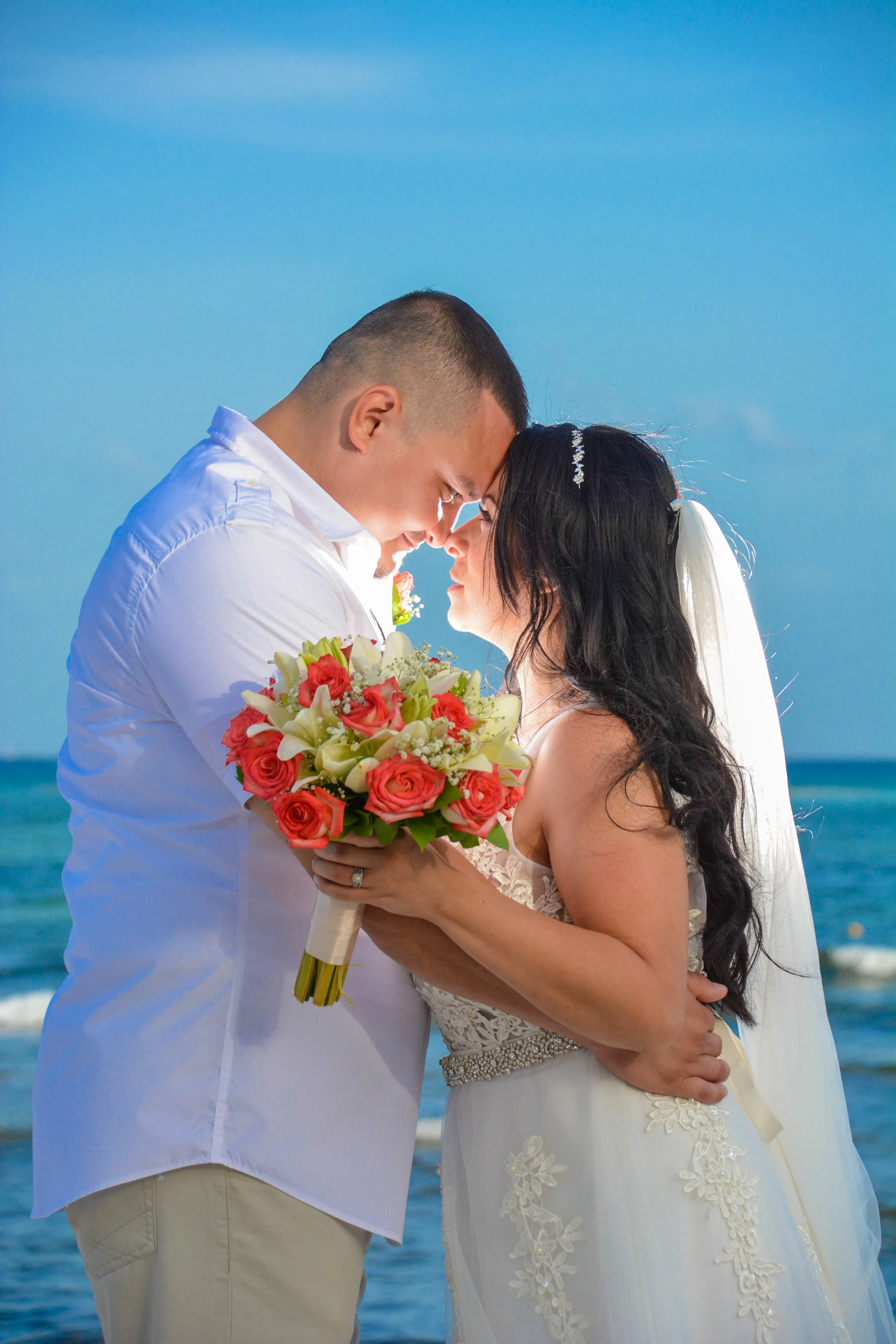 A bride and groom standing close on the beach, touching foreheads, holding a bouquet of pink roses, white lilies, and greenery, with the ocean and blue sky in the background.