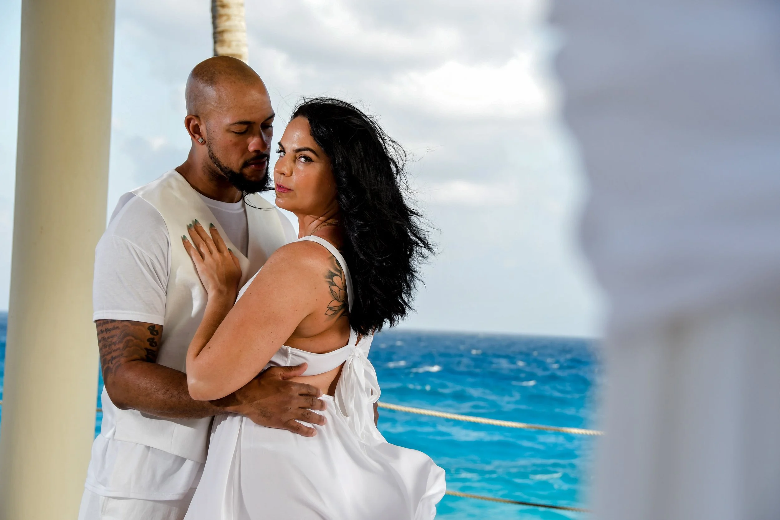 A man and woman embrace on a boat with the ocean in the background. The man has a shaved head, tattoos, and wears a white shirt. The woman has black hair, tattoos, and wears a white sleeveless dress. The sky is cloudy, and the scene is romantic.